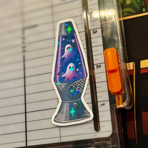 a star holographic sticker of a 70s style purple lava lamp with ghosts and bats floating inside the lamp. spider webs drawn on the base of the lamp. the sticker is placed on a Fiskars Paper Trimmer.