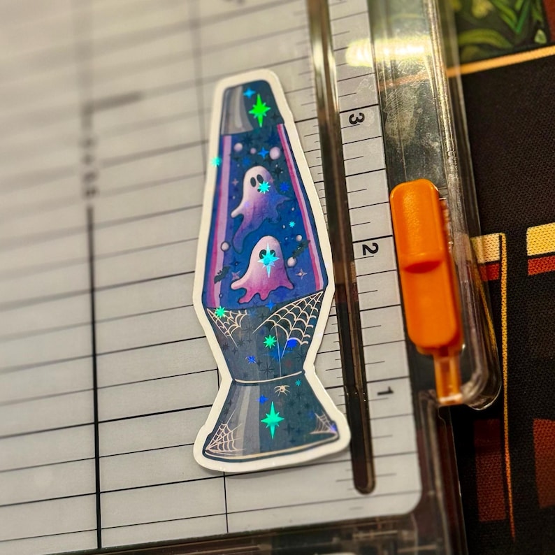 a star holographic sticker of a 70s style purple lava lamp with ghosts and bats floating inside the lamp. spider webs drawn on the base of the lamp. the sticker is placed on a Fiskars Paper Trimmer.