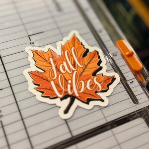 a sticker of a detailed orange fall leaf with the words in cursive Fall Vibes in the center. the sticker is placed on a Fiskars Paper Trimmer.