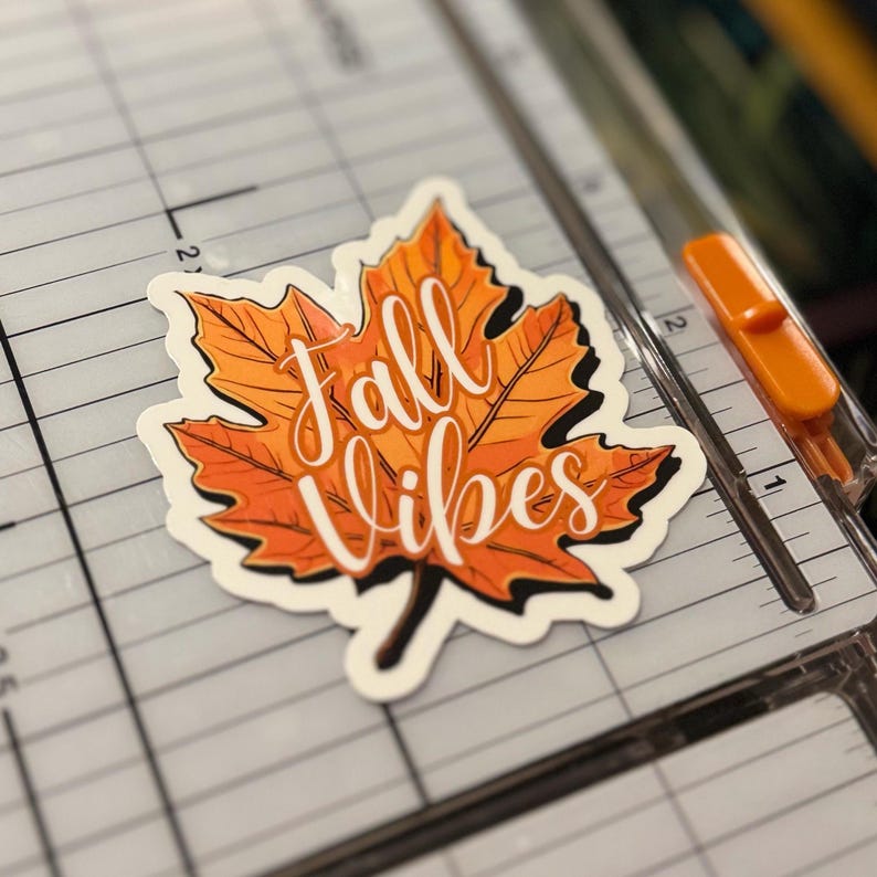 a sticker of a detailed orange fall leaf with the words in cursive Fall Vibes in the center. the sticker is placed on a Fiskars Paper Trimmer.