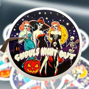 fine-point tweezers holding up a circle-shaped sticker with the words Ghouls Night Out and three ladies with sexy costumes and ghoul makeup on. Starry sky with moon, haunted house and dancing skeleton in background. duplicates on table in background.