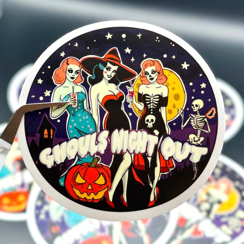 fine-point tweezers holding up a circle-shaped sticker with the words Ghouls Night Out and three ladies with sexy costumes and ghoul makeup on. Starry sky with moon, haunted house and dancing skeleton in background. duplicates on table in background.