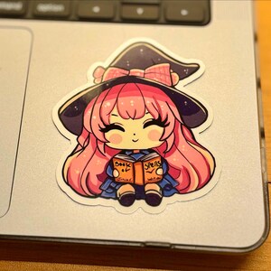 a sticker of a young witch pink hair. happy and smiling eyes closed, big black eyelashes. purple/black witch hat with pink bow. holding a book that says Book of Spells Junior Witch. the sticker is placed on a MAC laptop to the right of the trackpad.