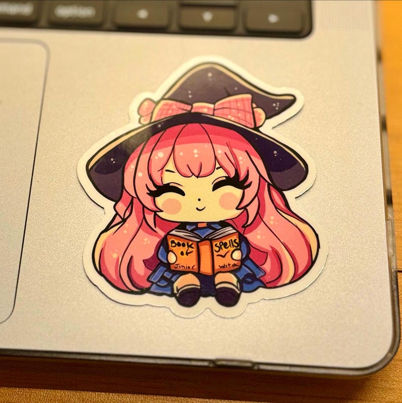 a sticker of a young witch pink hair. happy and smiling eyes closed, big black eyelashes. purple/black witch hat with pink bow. holding a book that says Book of Spells Junior Witch. the sticker is placed on a MAC laptop to the right of the trackpad.
