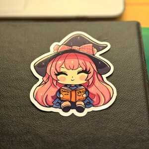 a sticker of a young witch pink hair. happy and smiling eyes closed, big black eyelashes. purple/black witch hat with pink bow. holding a book that says Book of Spells Junior Witch. the sticker is placed to the top right of a black 3-ring binder.