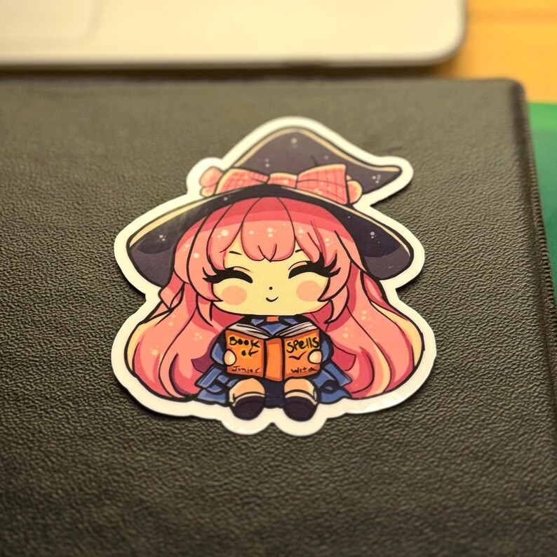 a sticker of a young witch pink hair. happy and smiling eyes closed, big black eyelashes. purple/black witch hat with pink bow. holding a book that says Book of Spells Junior Witch. the sticker is placed to the top right of a black 3-ring binder.