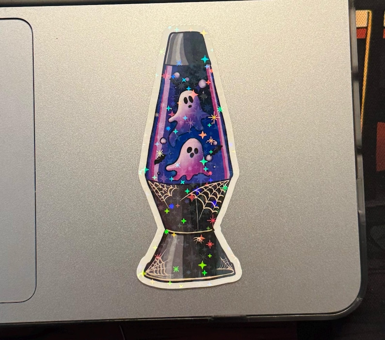 a star holographic sticker of a 70s style purple lava lamp with ghosts and bats floating inside the lamp. spider webs drawn on the base of the lamp. the sticker is placed on a MAC laptop to the right of the trackpad.