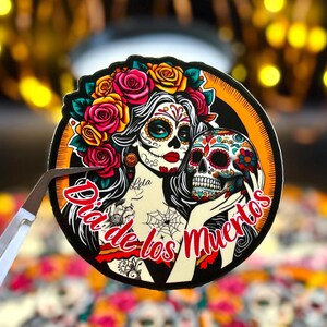 May include: A circular sticker featuring a woman with sugar skull makeup, wearing a crown of red, pink, and yellow roses. She holds a colorful decorated skull. The text Dia de los Muertos is written in red script across the bottom of the design.