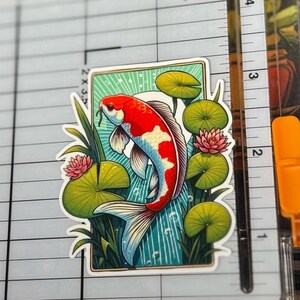 a rectangle shaped sticker of a koi fish swimming in a lilly pond, white sun and rays in background. the lillys stick out beyond the rectangle creating a 3D effect. the sticker is placed on a Fiskars Paper Trimmer.