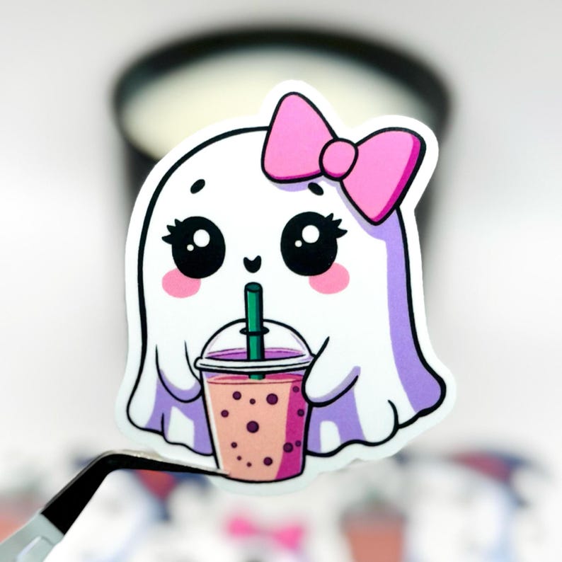 May include: A white ghost-shaped sticker with a pink bow, large eyes, and rosy cheeks. The ghost is drinking a pink bubble tea with a green straw. The sticker is held by a pair of tweezers, with other stickers in the background.