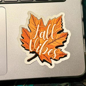 a sticker of a detailed orange fall leaf with the words in cursive Fall Vibes in the center. the sticker is placed on a MAC laptop to the right of the trackpad.