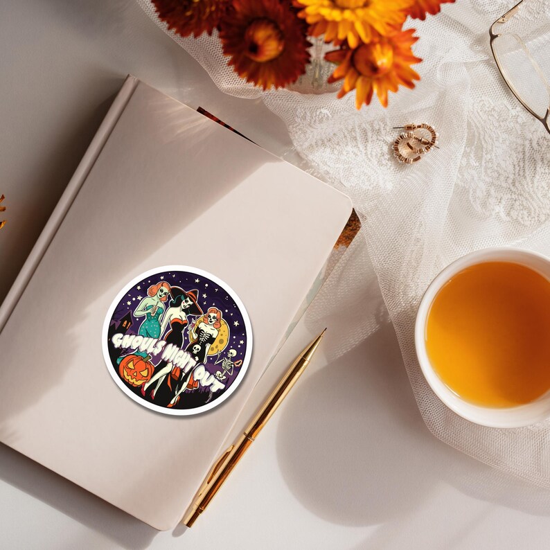 May include: A circular sticker featuring a dark purple night sky background with three stylized figures, a jack-o'-lantern, and a skeleton. The text GHOULS NIGHT OUT is printed in white across the center of the sticker, which is placed on a closed notebook.
