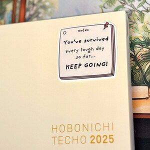 May include: A cream-colored Hobonichi Techo 2025 planner featuring a square sticker of a notepad with a red pushpin. The sticker text reads: notes, You've survived every tough day so far... KEEP GOING! The planner cover has gold text.