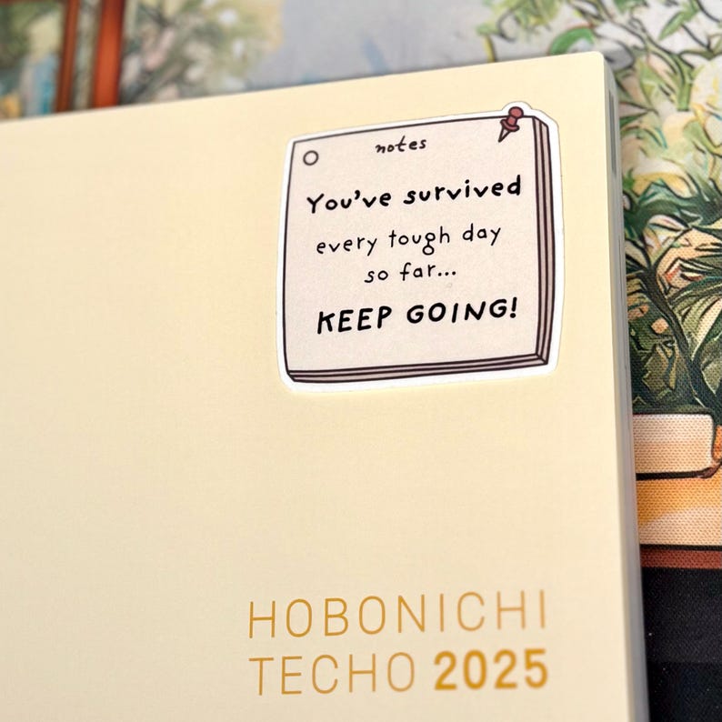 May include: A cream-colored Hobonichi Techo 2025 planner featuring a square sticker of a notepad with a red pushpin. The sticker text reads: notes, You've survived every tough day so far... KEEP GOING! The planner cover has gold text.