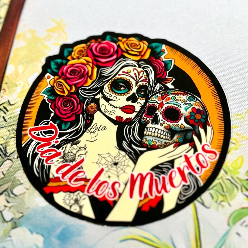 May include: A circular sticker featuring a woman with sugar skull face paint, wearing a crown of red and yellow roses, holding a decorated skull. The text Dia de los Muertos is written in red script across the bottom of the circular design.