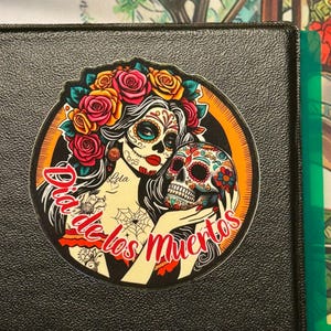 May include: A circular sticker featuring a woman with sugar skull makeup, a crown of red and yellow roses, and long dark hair. She holds a decorated skull in her hands. The text Dia de los Muertos is written in red script across the bottom.