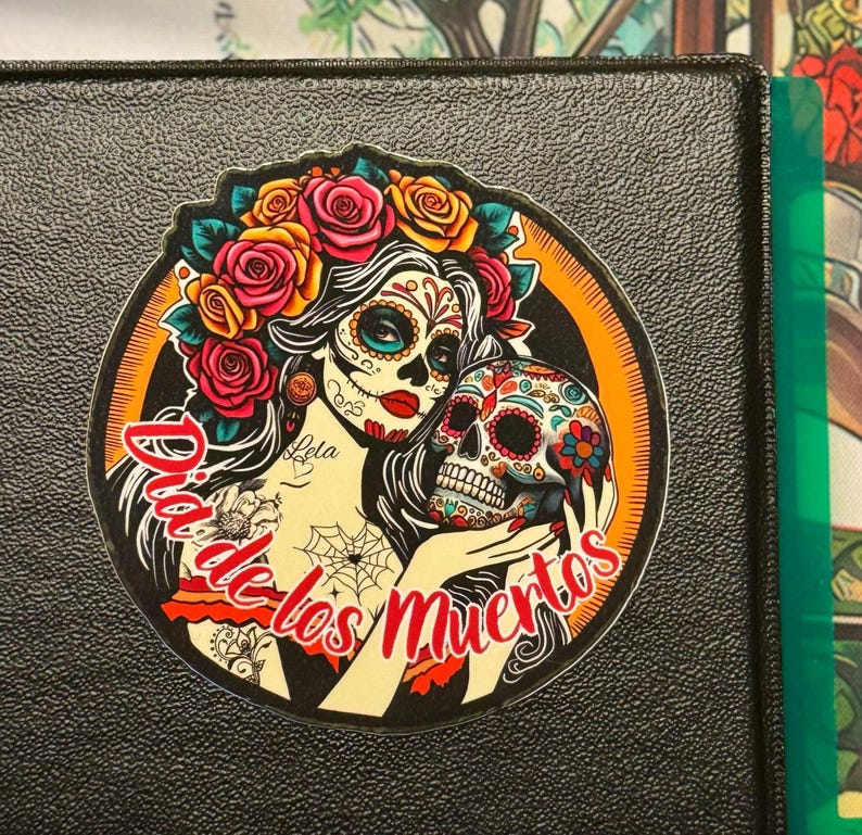 May include: A circular sticker featuring a woman with sugar skull makeup, a crown of red and yellow roses, and long dark hair. She holds a decorated skull in her hands. The text Dia de los Muertos is written in red script across the bottom.