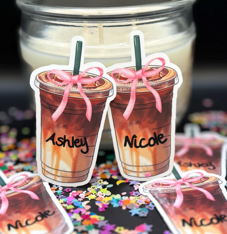 May include: Two iced coffee stickers with pink bows and names "Ashley" and "Nicole" written on them. The stickers are on a background of colorful confetti.