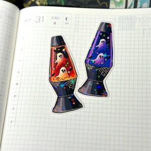 May include: Two holographic stickers shaped like lava lamps. One lamp features an orange liquid with white ghosts and black bats, while the other contains a blue and purple liquid with white ghosts and black bats. Both bases feature spider web designs.