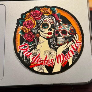 May include: A circular sticker featuring a person with sugar skull makeup, red lips, and a crown of red, orange, and yellow roses. The person holds a decorated skull. The text "Dia de los Muertos" is written in red script across the bottom.