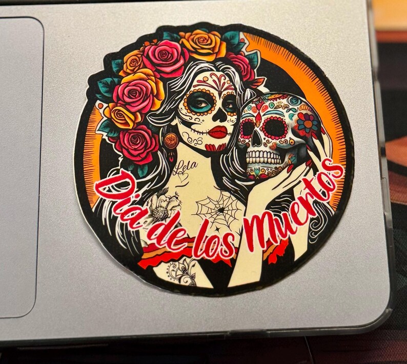May include: A circular sticker featuring a person with sugar skull makeup, red lips, and a crown of red, orange, and yellow roses. The person holds a decorated skull. The text "Dia de los Muertos" is written in red script across the bottom.