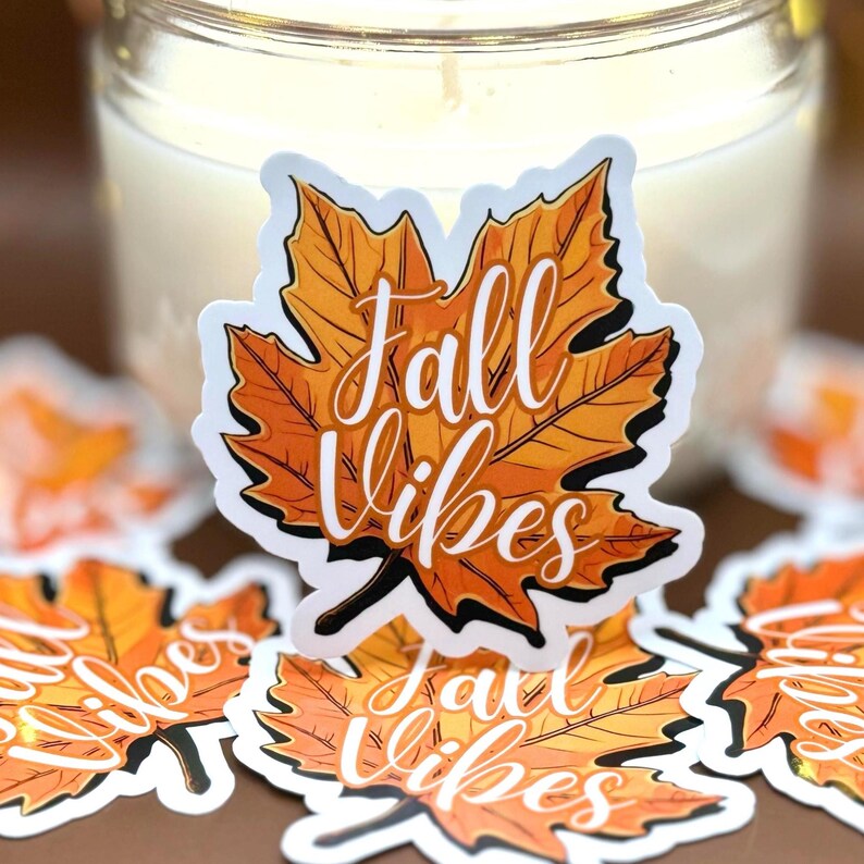a sticker of a detailed orange fall leaf with the words in cursive Fall Vibes in the center. the sticker is placed in front of a white candle, glowing string lights, and duplicates on the table in the background.