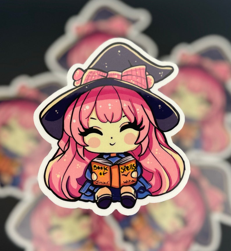a sticker of a young witch pink hair. happy and smiling eyes closed, big black eyelashes. purple/black witch hat with pink bow. holding a book that says Book of Spells Junior Witch. duplicates in blurred background.