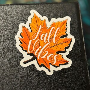 a sticker of a detailed orange fall leaf with the words in cursive Fall Vibes in the center. the sticker is placed to the top right corner of a black 3-ring binder.