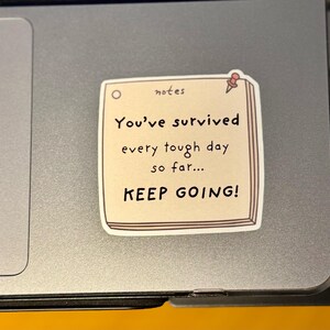 May include: A square sticker designed to look like a beige notepad with a red pushpin in the top right corner. The text reads: notes, You've survived every tough day so far... KEEP GOING! The sticker is applied to a smooth, metallic gray surface.