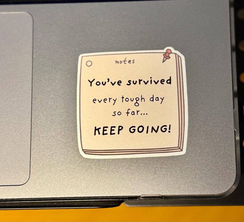 May include: A square sticker designed to look like a beige notepad with a red pushpin in the top right corner. The text reads: notes, You've survived every tough day so far... KEEP GOING! The sticker is applied to a smooth, metallic gray surface.