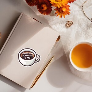 May include: A white sticker shaped like a coffee cup with dark brown liquid inside. The text "Maybe coffee is addicted To ME?" is printed in white on the liquid. The sticker is placed on a closed light-colored notebook next to a gold pen.