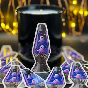 a star holographic sticker of a 70s style purple lava lamp with ghosts and bats floating inside the lamp. spider webs drawn on the base of the lamp. black candle, glowing string lights, and duplicates on the table in the background.