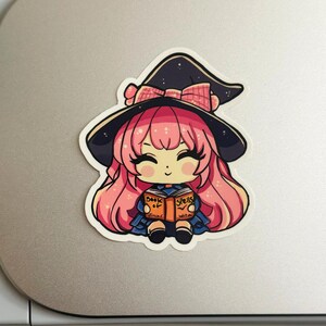 a sticker of a young witch pink hair. happy and smiling eyes closed, big black eyelashes. purple/black witch hat with pink bow. holding a book that says Book of Spells Junior Witch. the sticker is placed on a cricut cutting machine.