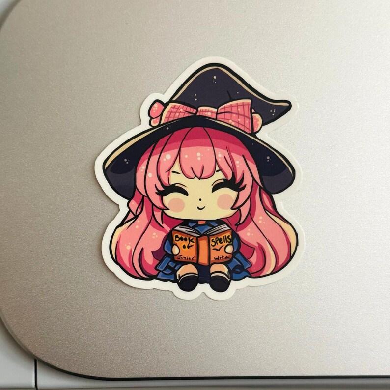 a sticker of a young witch pink hair. happy and smiling eyes closed, big black eyelashes. purple/black witch hat with pink bow. holding a book that says Book of Spells Junior Witch. the sticker is placed on a cricut cutting machine.