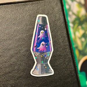 a star holographic sticker of a 70s style purple lava lamp with ghosts and bats floating inside the lamp. spider webs drawn on the base of the lamp. the sticker is placed to the top right of a black 3-ring binder.