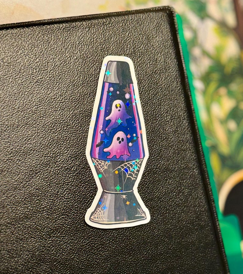 a star holographic sticker of a 70s style purple lava lamp with ghosts and bats floating inside the lamp. spider webs drawn on the base of the lamp. the sticker is placed to the top right of a black 3-ring binder.