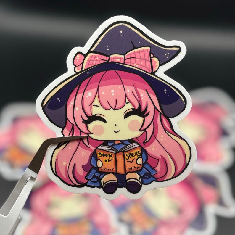 fine-point tweezers holding a sticker of a young witch pink hair. happy and smiling eyes closed, big black eyelashes. purple/black witch hat with pink bow. holding a book that says Book of Spells Junior Witch on the front. duplicates in background.