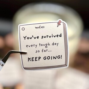 May include: A square sticker designed to look like a notepad page with a light beige background and a brown border. It features a small red pushpin icon and the text: notes, You've survived every tough day so far... KEEP GOING!