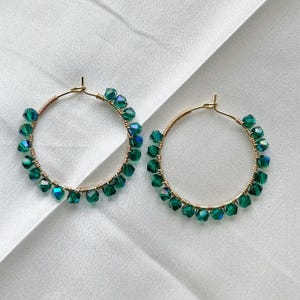 May include: A pair of gold hoop earrings featuring faceted emerald green crystal beads wrapped around the wire. The circular earrings have a metallic gold finish and are adorned with multiple small, sparkling green beads.