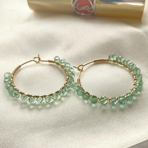 Può includere: Un paio di orecchini a cerchio dorati adornati con piccole perline verde chiaro. Le perline sono infilate lungo la metà inferiore dei cerchi, creando un design delicato ed elegante. Gli orecchini sono su uno sfondo bianco e setoso.