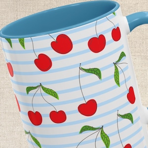 May include: White ceramic mug with a blue interior and handle. The mug features a pattern of red cherries with green leaves and thin blue stripes. The mug is designed for hot or cold beverages.