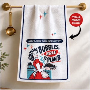 May include: A white waffle-weave kitchen towel with a dark blue border. The graphic features a retro-style illustration of a person in a red dress holding a platter, with text reading LEENA'S DINNER PARTY EMERGENCY KIT, BUBBLES, BRIE & PLAN B.