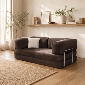 May include: A brown corduroy sofa with a white square pillow. The sofa has a low profile and a minimalist design. The room features a neutral color palette with a woven rug and wooden accents.