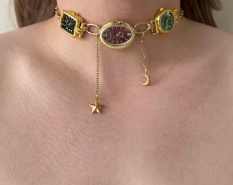 May include: A gold-toned choker necklace featuring three vintage watch faces in black, burgundy, and green. The necklace has dangling chains with a gold star and crescent moon charm. The watch faces are rectangular and oval.