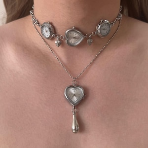 May include: A silver necklace with a choker featuring three small watch faces and heart charms. A longer chain holds a heart-shaped watch pendant with a teardrop charm.