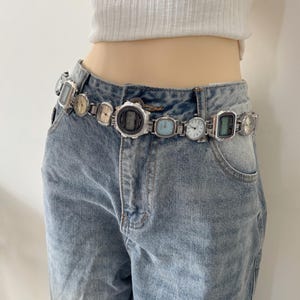 Könnte beinhalten: Ein Gürtel aus einer Reihe miteinander verbundener silberfarbener Armbanduhren, bestehend aus einer Mischung von analogen Zifferblättern in Weiß oder Hellblau und digitalen Anzeigen mit schwarzen Bildschirmen, verbunden durch Metallarmbänder.