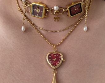 May include: A gold-toned necklace featuring three square-faced watches with burgundy dials, connected by pearl accents and a bow. A heart-shaped watch pendant with a burgundy face and a teardrop charm hangs below. The necklace is a unique jewelry piece.