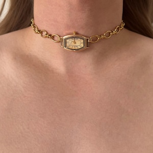 Puede incluir: Un collar gargantilla con reloj dorado. La esfera del reloj es rectangular con números romanos y agujas doradas. La cadena está hecha de eslabones grandes y ovalados. El collar se lleva alrededor del cuello.