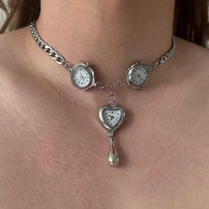 Può includere: Collana color argento con tre quadranti di orologio. Due quadranti rotondi sono sulla catena e un quadrante a forma di cuore con un pendente a goccia è appeso al centro. Gli orologi hanno quadranti bianchi e numeri neri.