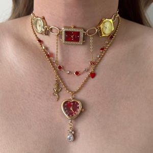 May include: A gold-toned necklace featuring multiple watch faces and decorative elements. The necklace includes a rectangular watch with a red face, heart-shaped watch pendant, and red and clear gem accents. The design incorporates a rose and a dangling teardrop-shaped gem.